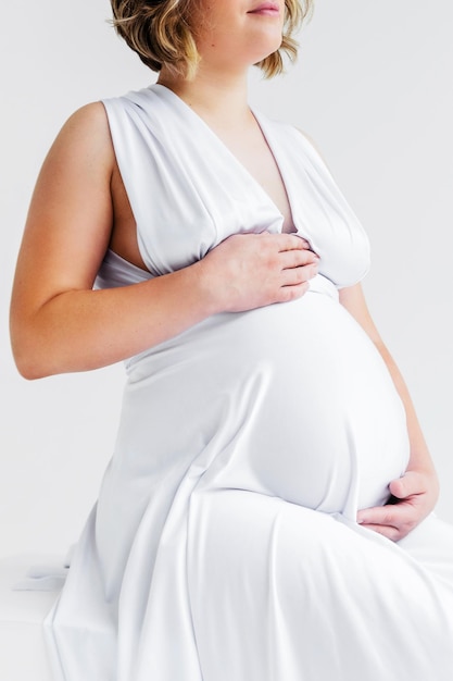 A pregnant woman in a long white silk dress hugs her belly and poses on a white background