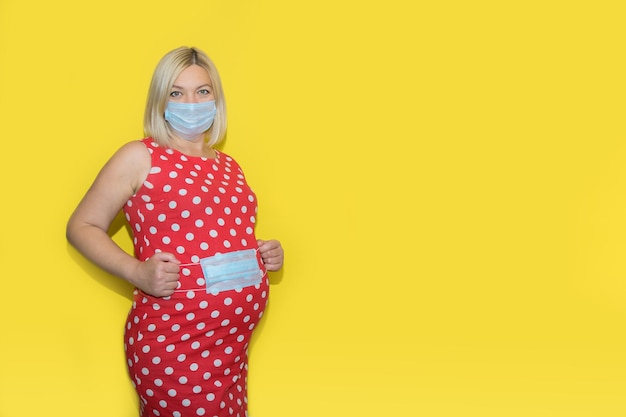 Pregnant woman in a mask holds a mask in her hands covering her belly on a yellow background with copy space