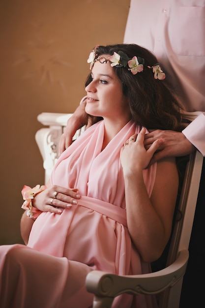 Pregnant woman in a pink dress sitting in a chair behind a smiling man standing. expecting a baby.