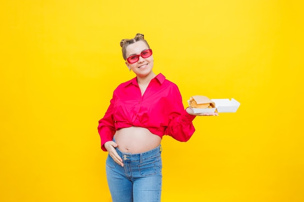 A pregnant woman in a pink shirt and jeans is holding a tasty burger Harmful food during pregnancy Concept of easy and happy pregnancy