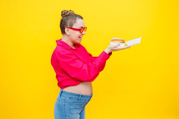 A pregnant woman in a pink shirt and jeans is holding a tasty burger Harmful food during pregnancy Concept of easy and happy pregnancy