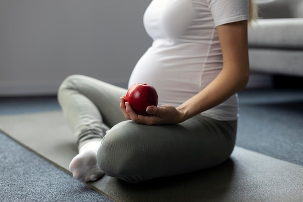 Pregnant woman practicing yoga, holding red apple at home. Pregnancy, healthy lifestyle concept