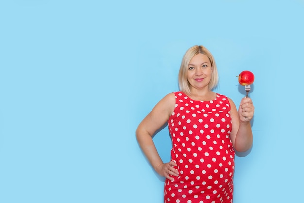 Pregnant woman in a red polka dot dress holding an apple on a fork on a blue background with copy space