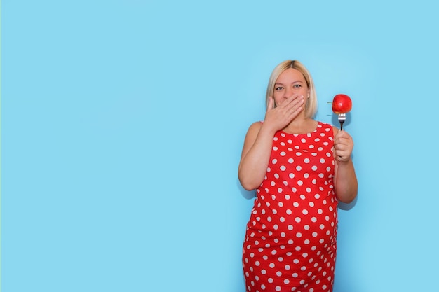 Pregnant woman in a red polka dot dress with an apple on a fork in her hand closes her mouth with the other hand on a blue background with copyspace the concept of nutrition for pregnant women