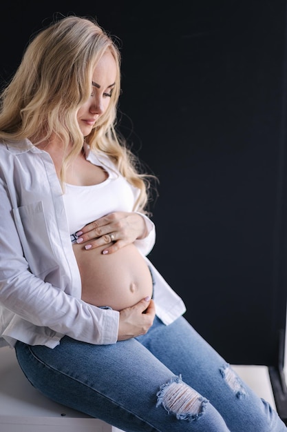 Pregnant woman sits on white cube an put hands on belly denim style white shirt and jeans blond hair