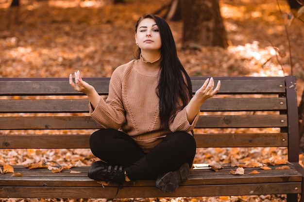 Pregnant woman sitting on the bench in autumn city park forest, meditating with her round belly