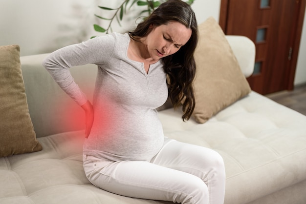 Pregnant woman sitting on couch with hand on back experiencing pain in back, backache concept, beginning of childbirth