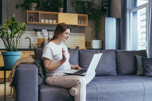 Pregnant woman sitting at home on the couch with a laptop talking on a video call to the doctor consults waves greets smiles