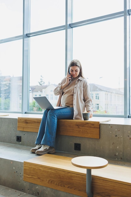 Pregnant woman sitting with laptop smartphone looking at laptop and taking notes while sitting in cafe A woman works remotely on a laptop while on maternity leave