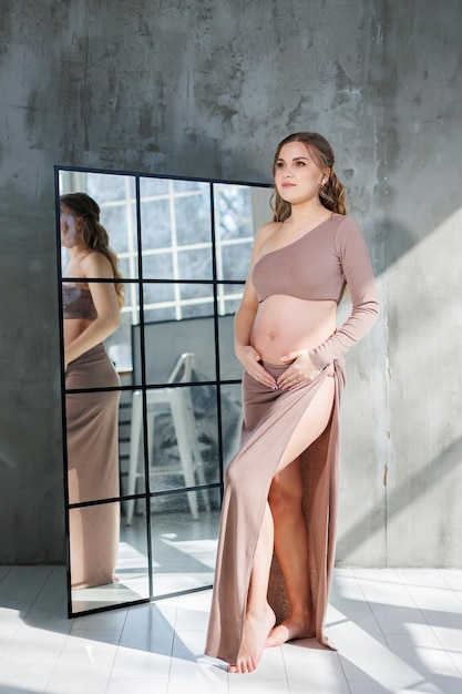 A pregnant woman in a skirt hugs her stomach with her hands Fashion pregnant woman Full length portrait on gray background Happy pregnancy