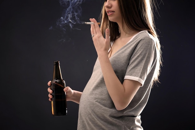 Pregnant woman smoking cigarette and drinking beer on dark background