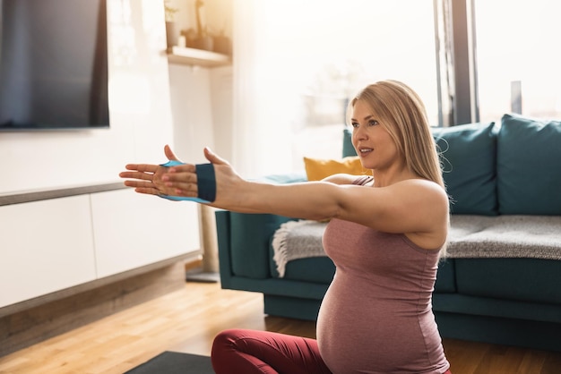 A pregnant woman in sports clothing is stretching with rubber band and exercising to promote wellbeing in her living room.