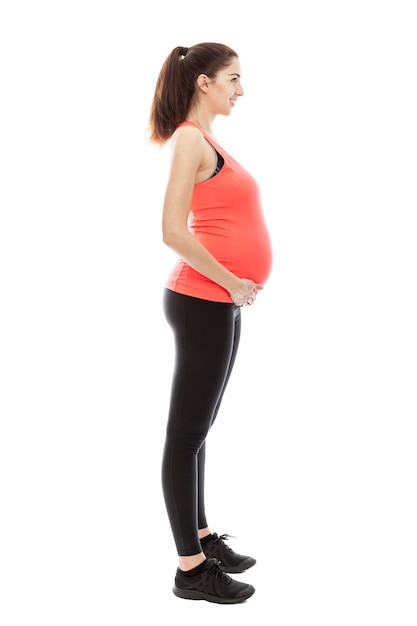 A pregnant woman in sportswear is standing in full growth fashioning. Profile view. Isolated on white wall. Vertical.