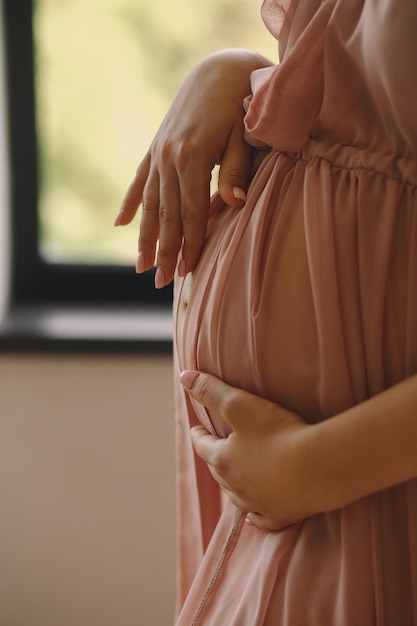 Photo pregnant woman in a stylish pink dress standing near window and touching her belly indoor