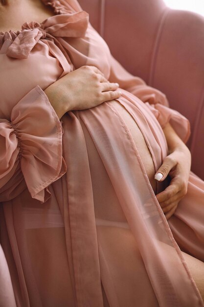 Photo pregnant woman in a stylish pink peignoir sitting on the chair and holding her belly pregnant belly