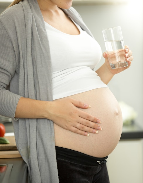 Pregnant woman on third trimester posing with glass of water on kitchen