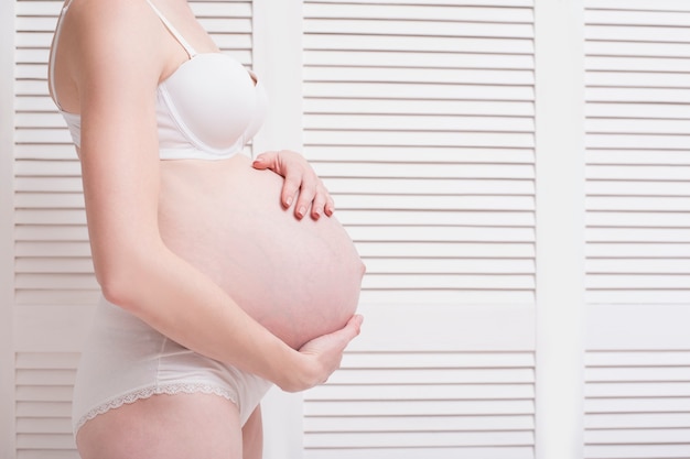 Pregnant woman in underwear hugging belly with hands on a light background.