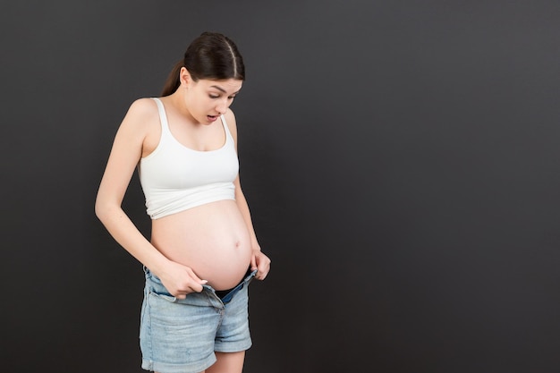 Pregnant woman in unzipped jeans showing her naked abdomen at colorful background with copy space