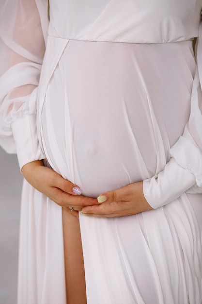 Photo pregnant woman in white dress holding her belly indoor close up photo of pregnant belly