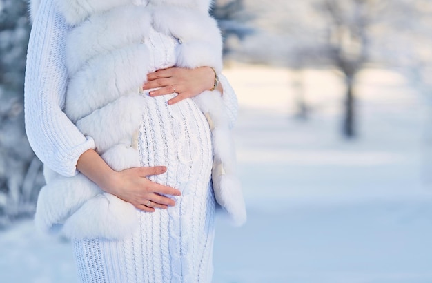 A pregnant woman in a white knitted dress and a fur vest in the forest in winter