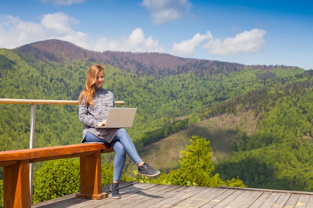 Pregnant woman with beautiful hair is searching something in her laptop sitiing on a bench on the background of nature