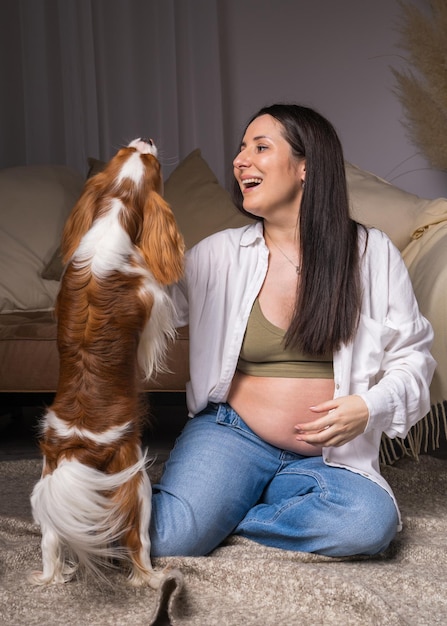 pregnant woman with exposed belly sits on the floor at home laughs happily and plays with her dog