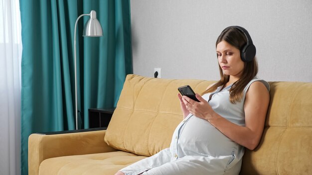 Pregnant woman with headphones looking at the phone while sitting on the couch.