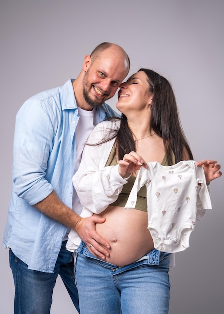 A pregnant woman with her belly exposed next to her husband holding baby clothes in her hands