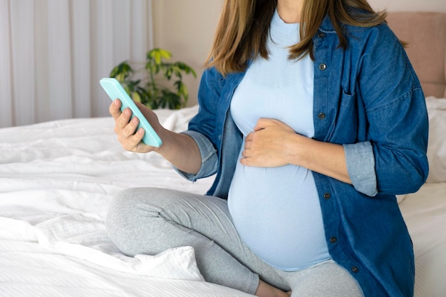 Pregnant woman with smartphone sitting on bed at home using pregnancy app chatting online surfing social media or watching video Maternity and technology