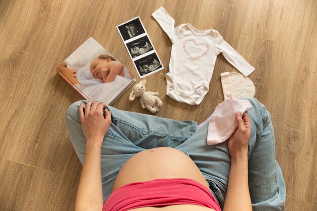 Pregnant woman with white baby bodysuit, socks, hat ultrasound image preparing to childbirth during pregnancy. Young mother sitting on the floor