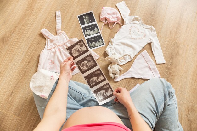 Pregnant woman with white baby bodysuit, socks, hat ultrasound image preparing to childbirth during pregnancy. Young mother sitting on the floor