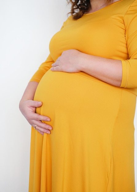 A pregnant woman in a yellow dress strokes her belly and poses on a white background Closeup of the belly