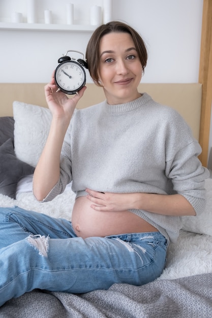 Pregnant young adult woman with alarm clock on bed at home, giving birth soon concept