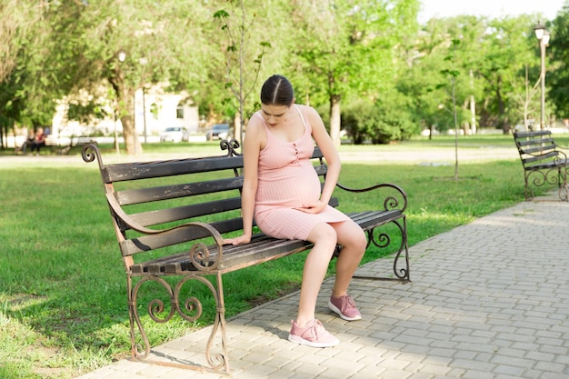 A pregnant young woman on a bench sitting in the park and holds on to the stomach which hurts Pregnancy and motherhood back pain during pregnancy