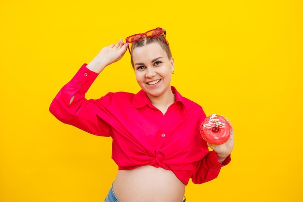 Pregnant young woman in a pink shirt with a donut in her hands Junk food during pregnancy Hungry pregnant woman holding donuts while standing on yellow background