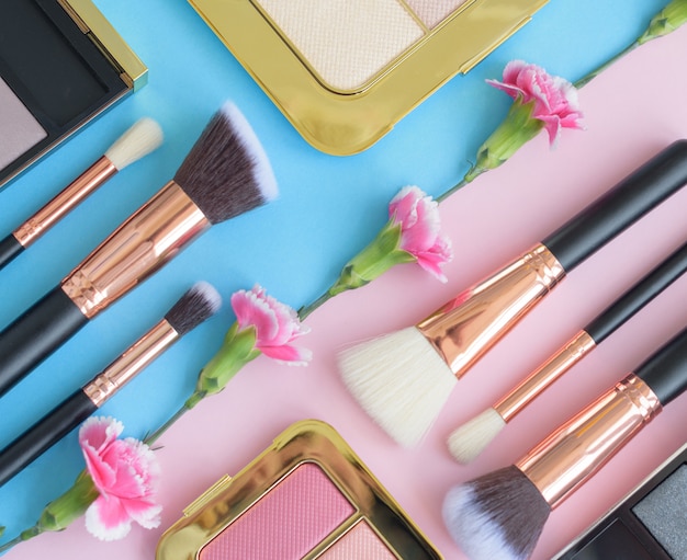 Premium makeup brushes, eye shadow palette and flowers on a colored blue and pink background, creative cosmetics flat lay with diagonal composition