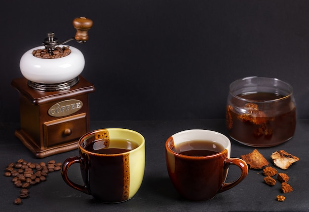 Preparation Chaga mushrooms coffee. Two two-colors ceramic cups, glass jar with chaga drink, coffee grinder on black.