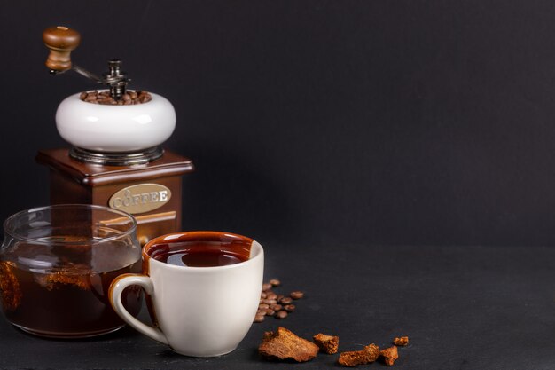 Preparation Chaga mushrooms coffee. White-brown cup and glass jar of chaga drink, coffee grinder on black background.