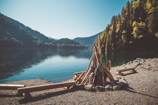 Photo prepared for kindling a large bonfire and benches from the logs on the shore of a beautiful lake with clear water surrounded by mountains and forest. toned image