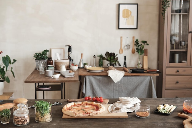 Prepared pizza on wooden board placed on table with ingredients and jars of spices table with utensils and crockery in background