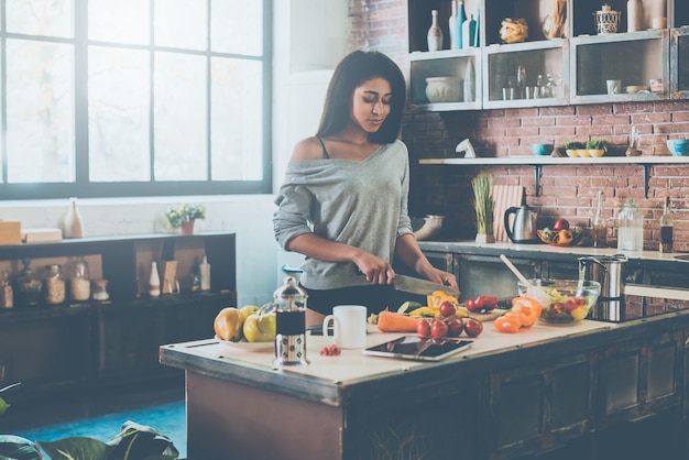 Preparing healthy food. Beautiful young mixed race woman cutting vegetables for salad while standing in kitchen at home