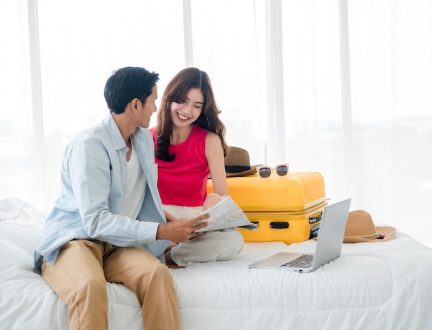 Preparing trip and vacation plan concepts Young pretty Asian woman and man in denim shirt looking at the laptop and map on the bed with suitcase in bedroom Travel planning on summer holiday