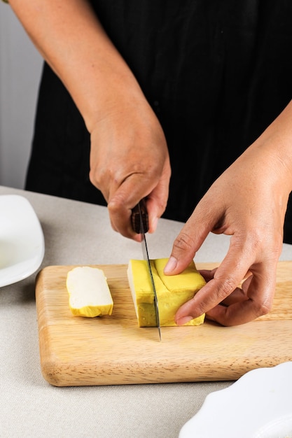 Preparing Vegan Meal. Female Asian Chefâs Hand Cutting Yellow Tofu with Knife on Wooden Board