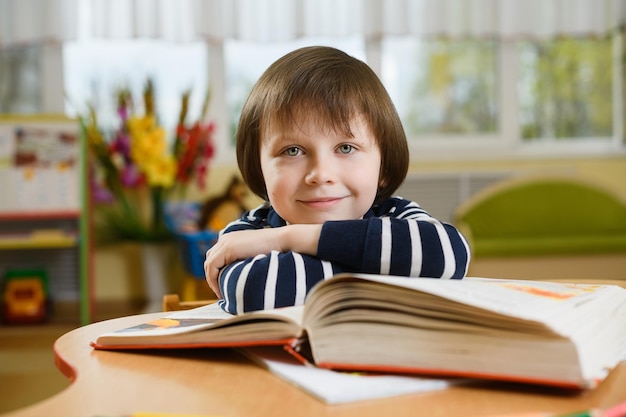 Preschool boy leaning his elbows on large book and smiles looking forward