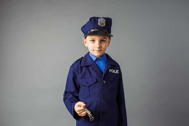 The preschool boy in a suit of a policeman with handcuffs stands on a gray background