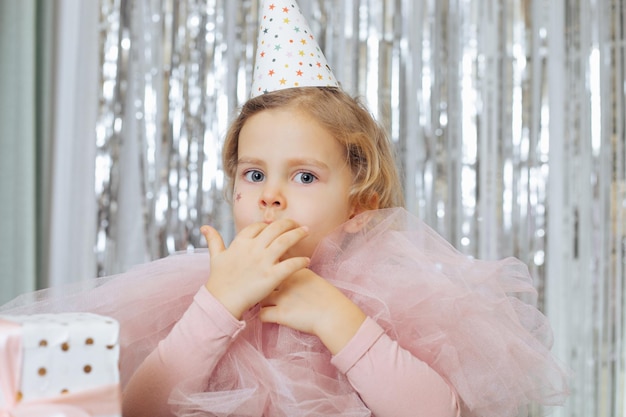 Preschool fair-haired girl having festival pink dress, party hat, stars decoration on her face, licking fingers with pleasure indoors possibly in cafe with decorated background of silvery spangle