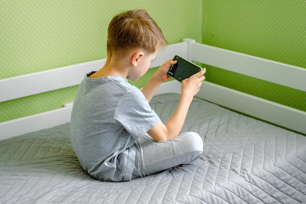 A preschool generation z boy sitts on the bed and plays a game on mobile Kids and technology