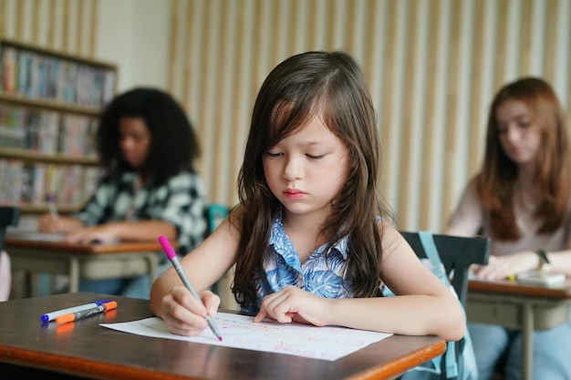 Preschool Girl Kid Drawing With Color Pencil On White Paper On Table In Classroom With Friends