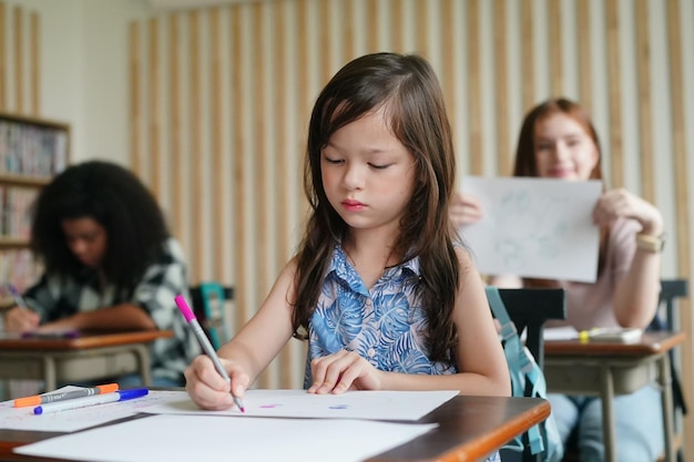 Preschool Girl Kid Drawing With Color Pencil On White Paper On Table In Classroom With Friends