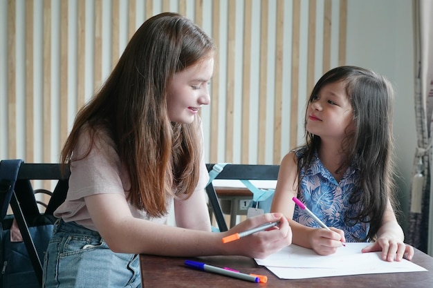 Preschool Girl Kid Drawing With Color Pencil On White Paper On Table In Classroom With Friends